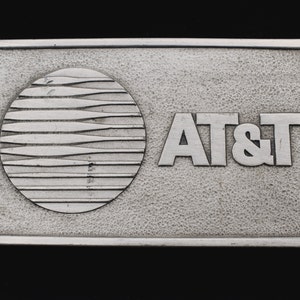 May include: A rectangular, silver-colored belt buckle with the AT&T logo. The logo features a stylized globe with horizontal lines and the letters "AT&T" in a bold, sans-serif font. The buckle has a textured background.
