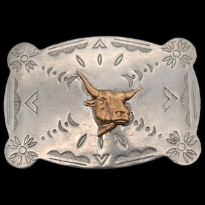 May include: Silver-toned rectangular belt buckle with a gold-colored bull head in the center. The buckle has etched floral and geometric designs around the edges. A classic Western-style accessory.