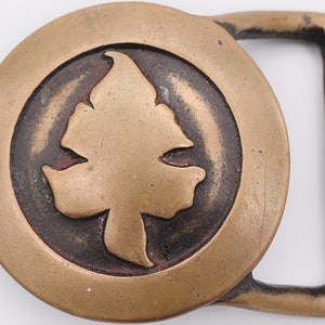 May include: A brass belt buckle with a stylized leaf design. The buckle is circular and has a raised edge. The leaf is in the center of the buckle and is raised.