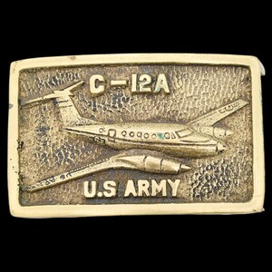 May include: A gold-tone metal belt buckle featuring a detailed image of a C-12A aircraft with the text "C-12A" and "U.S. ARMY" on the buckle.