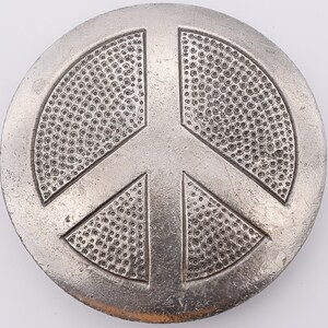 May include: A round, silver-colored peace symbol with a textured surface. The peace sign is a central design element, with a textured pattern filling each section. The symbol is set against a white background.