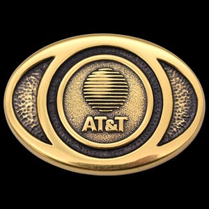 May include: A gold-colored oval belt buckle with the AT&T logo in the center. The logo features a stylized globe with a circle of lines around it. The text "AT&T" is below the globe.