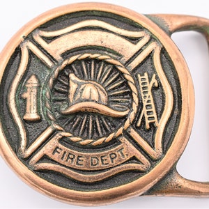 May include: Bronze-colored belt buckle with a firefighter theme. The circular buckle features a fire helmet, ladder, and hydrant, with the words "FIRE DEPT." The buckle has a rectangular loop and is made of metal.
