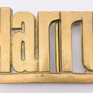 May include: A rectangular brass belt buckle with the name "harry" cut out in large block letters. The buckle has a gold finish with some signs of wear and a brass bar for the belt.