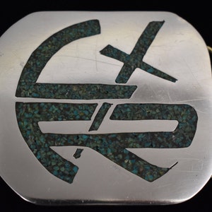 May include: Silver belt buckle with a turquoise inlay design. The design is a stylized geometric pattern.
