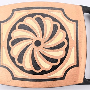 May include: A rectangular belt buckle with a copper-colored base and a black handle. The center features a floral design with black, cream, and copper-colored accents. The buckle is approximately 3 inches wide and 2 inches tall.