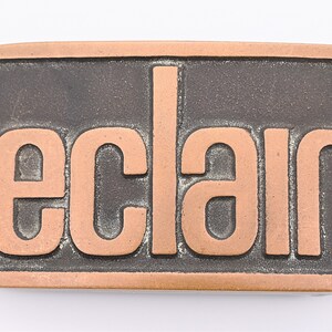 May include: Rectangular belt buckle with the word "eclair" in copper-colored raised letters against a dark gray background. The buckle has a copper-colored border and a vintage aesthetic.