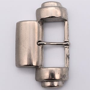 May include: A silver-toned metal belt buckle with a rectangular frame and a cylindrical post. The buckle has a classic design, suitable for various belts. The metal has a slightly aged appearance.
