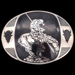 May include: Oval silver-tone belt buckle with a black center featuring a Native American warrior on horseback holding a spear. Two bison silhouettes flank the central design. The buckle has a decorative border.