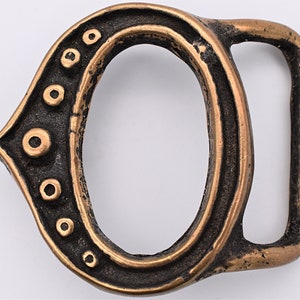 May include: A bronze-colored metal belt buckle with an oval shape and a rectangular bar. The buckle has a decorative design with raised circular accents along one side, and a dark patina.