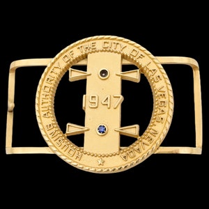 May include: A gold-colored belt buckle with the text "HOUSING AUTHORITY OF THE CITY OF LAS VEGAS NEVADA" encircling a central design. The year "1947" is displayed prominently. The buckle features two small, dark gemstones and one blue gemstone.