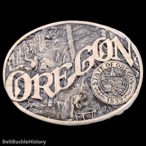 May include: An oval belt buckle featuring a relief design of the Oregon landscape with the word OREGON in large letters and the State of Oregon seal with the text STATE OF OREGON 1859. The metallic surface shows a forest scene with figures.