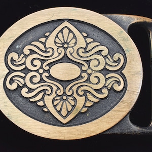 May include: A vintage brass belt buckle with an ornate, symmetrical design. The central oval is surrounded by swirling patterns and floral elements, set against a dark background. The buckle has a rectangular frame and a brass finish.