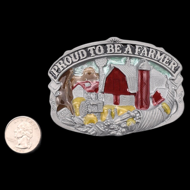 May include: Silver-toned belt buckle with a farm scene and the text "PROUD TO BE A FARMER". The buckle features a red barn, a silo, and a tractor. A U.S. quarter is next to the buckle for size comparison.