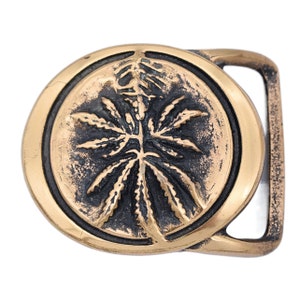 May include: A gold-colored metal belt buckle with a circular design featuring a cannabis leaf. The leaf is dark brown, set against a lighter gold background. The buckle has a rectangular frame and a classic pin closure.