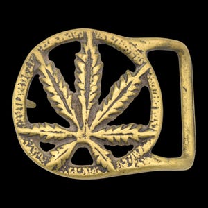 May include: A brass-colored belt buckle featuring a cannabis leaf design. The leaf is centered within a circular frame, with the buckle's rectangular prong on the right side. The buckle has a textured, aged appearance against a black background.