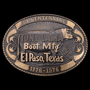 May include: Oval-shaped, bronze-toned belt buckle with the text "BI-CENTENNIAL" and "TONY LAMA Boot Mfg El Paso, Texas 1776-1976". A bull graphic is in the center. The buckle has a rope-like border.