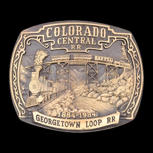 May include: A detailed, oval-shaped belt buckle with a train scene. The buckle features the text "COLORADO CENTRAL RR" and "GEORGETOWN LOOP RR" with the years "1884-1984". The design includes a train on a bridge and a train on the tracks.