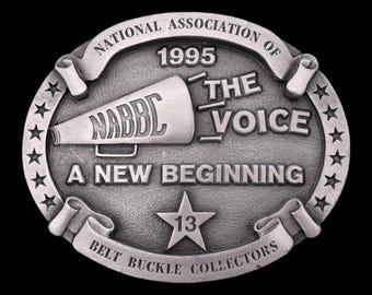 May include: Oval silver-colored belt buckle with the text "NATIONAL ASSOCIATION OF BELT BUCKLE COLLECTORS 1995" and "THE VOICE A NEW BEGINNING." Features a megaphone, stars, and a star with the number 13. The buckle has a detailed design.