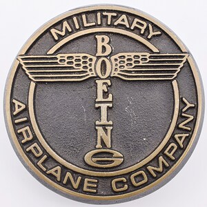 May include: A round, bronze-colored metal emblem with the words "MILITARY AIRPLANE COMPANY" encircling the edge. The word "BOEING" is in the center, with stylized wings above and a stylized "G" below.