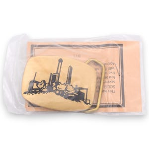 May include: A gold-colored metal belt buckle with a black silhouette design of a cityscape. The buckle is rectangular with rounded edges and a brass-colored clasp. The buckle is packaged in a clear plastic bag.