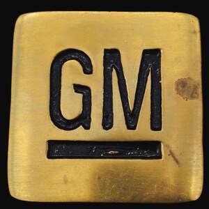 May include: A square, gold-colored metal object with the black letters "GM" and a black horizontal line. The object has a slightly worn appearance and is set against a black background.