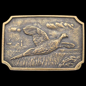 May include: A rectangular, antique-style belt buckle with a detailed relief of a pheasant in flight over a field. The buckle has a gold-toned finish and a decorative border, with a rural landscape background.