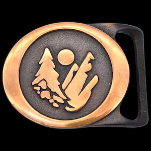 May include: Oval metal belt buckle with a nature scene. The buckle features a bronze-colored outer ring and a dark gray center with a mountain, tree, and sun design in bronze. The buckle has a black bar for the belt.
