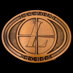 May include: Bronze-colored oval belt buckle with the words "LEUPOLD" and "SIGHTS" around a central circular design. The design features a crosshair and the letter "L".
