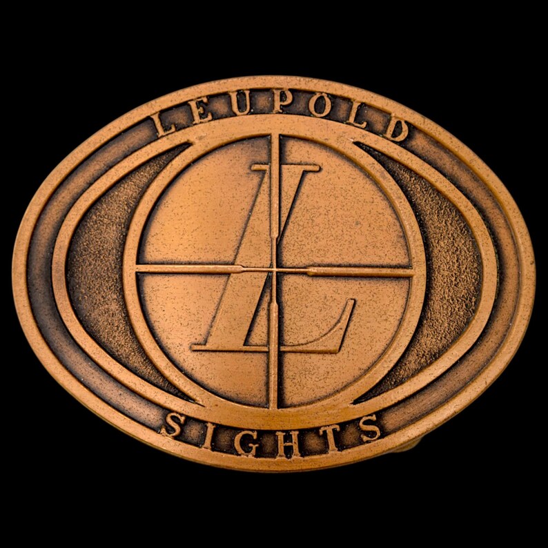 May include: Bronze-colored oval belt buckle with the words "LEUPOLD" and "SIGHTS" around a central circular design. The design features a crosshair and the letter "L".