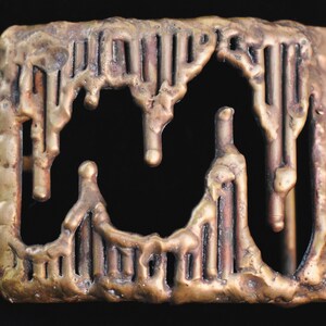 May include: A rectangular, bronze-colored belt buckle with a textured, melted appearance. The buckle features vertical and horizontal bars with a dripping design, creating an abstract, artistic look. The buckle is set against a black background.