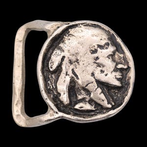 May include: A silver-colored belt buckle featuring a detailed profile of a Native American chief. The buckle has a circular design with a rectangular loop for the belt. The background is black.