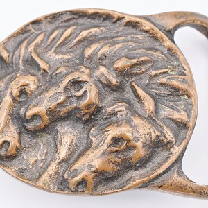 May include: Bronze-colored belt buckle featuring a detailed relief of three horse heads. The buckle has a rounded shape with a rectangular bar for the belt. The horses' manes and faces are intricately sculpted.