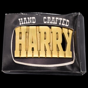 May include: A gold-colored metal belt buckle with the name "HARRY" in bold letters. The buckle is set against a black background and is labeled "HAND CRAFTED" in white text.