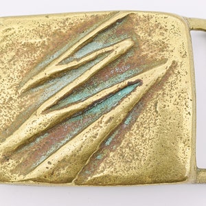 May include: A brass belt buckle with a textured, abstract design. The buckle has a greenish patina and a rectangular shape.