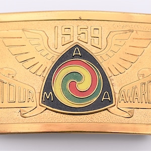 May include: Gold-colored metal belt buckle with a black, yellow, red, and green swirl design inside a triangle. The triangle has the letters "AMA" on the points and the year "1959" above. The buckle also has two wings and a scroll that reads "Tour Award".