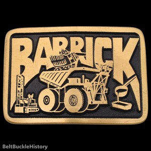 May include: A rectangular brass-toned belt buckle featuring a black recessed background. The word BARRICK is embossed in large letters across the top. Below the text, a mining dump truck and industrial equipment are depicted in relief.