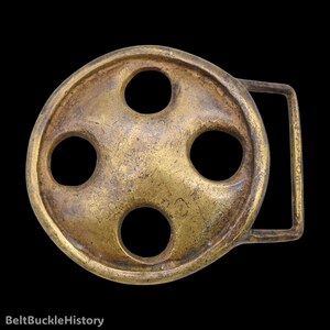 May include: A circular brass belt buckle with a weathered, golden-brown patina. The design features four evenly spaced circular cutouts arranged around a central point, connected to a rectangular loop on the right side for belt attachment.