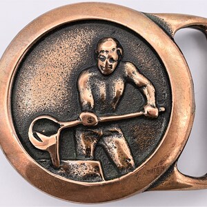 May include: A copper-colored belt buckle with a circular design. The center features a relief of a blacksmith working with a tool. The buckle has a rectangular bar for the belt and a circular frame around the blacksmith image.