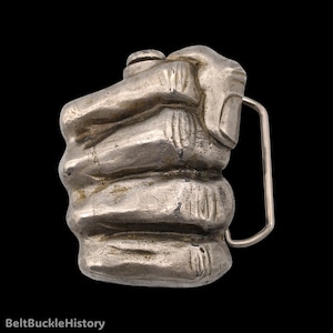 May include: A silver-toned metal belt buckle shaped like a clenched fist. The design features detailed finger joints and knuckles with a weathered, metallic finish. A rectangular metal loop is attached to the right side for belt attachment.