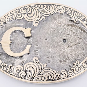 May include: A silver-toned belt buckle with an ornate design and the letter "C" in the center.