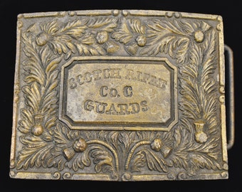 May include: A rectangular, antique brass belt buckle with the text "SCOTCH RIFLE Co. C GUARDS" in the center. The buckle is framed by ornate, embossed foliage and thistle designs, with a standard belt loop on the right side.