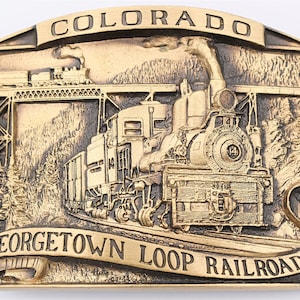May include: A gold-toned metal belt buckle featuring a detailed relief sculpture of a steam locomotive traveling through a mountain pass. The buckle is inscribed with "COLORADO GEORGETOWN LOOP RAILROAD".