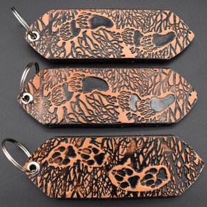 May include: Three brown leather keychains with silver key rings. Each keychain has a black and brown design of paw prints and branches.