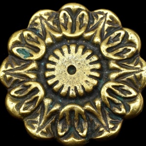 May include: A gold-toned metal decorative button with a circular design featuring a central wheel-like motif surrounded by a ring of stylized leaves.