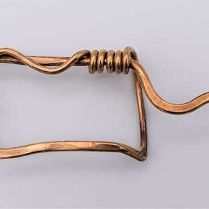 May include: A copper belt buckle with a unique, twisted design. The buckle is rectangular with a rounded edge and a small, protruding pin on the side. The buckle is made of a single piece of copper that is bent and twisted to create the design.