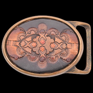 May include: Oval-shaped belt buckle with a detailed, embossed design. The central area features a dark brown background with a copper-colored floral pattern. The buckle has a bronze-colored frame and a matching clasp, set against a black background.