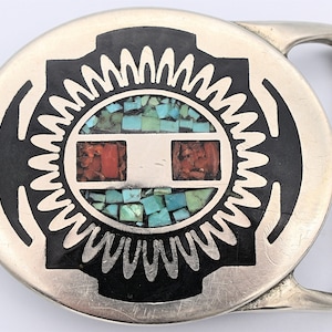 May include: A silver-toned belt buckle with a circular design. The center features a mosaic of turquoise and coral-colored stones. A black, stylized sunburst pattern surrounds the center. The buckle is rectangular with a curved bar.