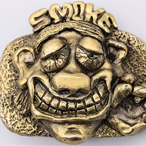 May include: A gold-tone metal belt buckle featuring a cartoon face with a wide grin and the word "SMOKE" above the head.