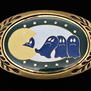 May include: A gold-tone belt buckle with a Pac-Man design. The buckle features a yellow Pac-Man eating three blue ghosts with white eyes.